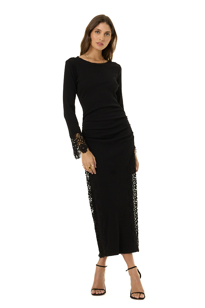A woman modeling a black long-sleeve midi dress with sheer lace side panels and lace-trimmed cuffs, paired with black strappy high-heeled sandals against a plain white background.