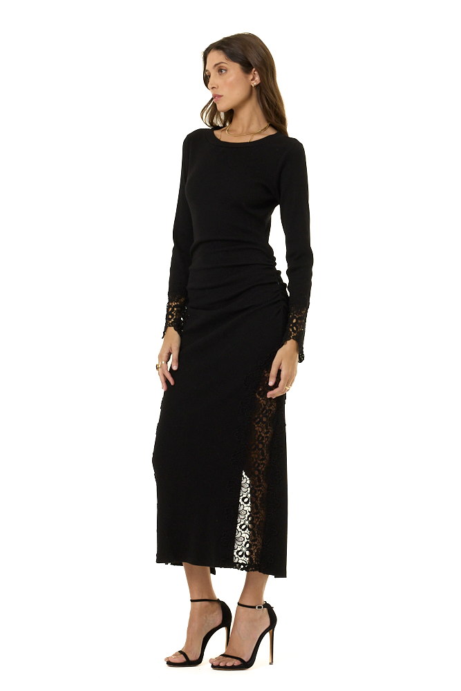 A woman modeling a black long-sleeve midi dress with sheer lace panels on the sides and cuffs, paired with black high-heeled sandals, against a plain white background.