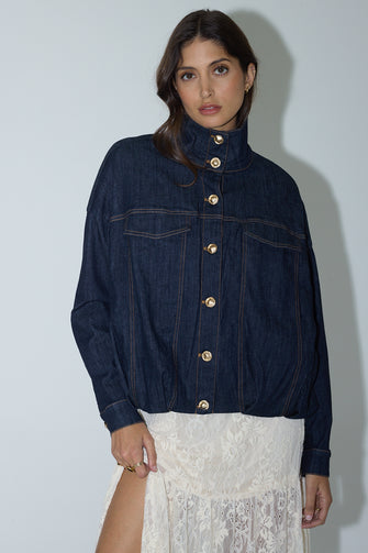 A woman wearing a dark indigo denim jacket with gold-tone buttons and a high collar, paired with a white lace skirt, standing against a light gray wall.