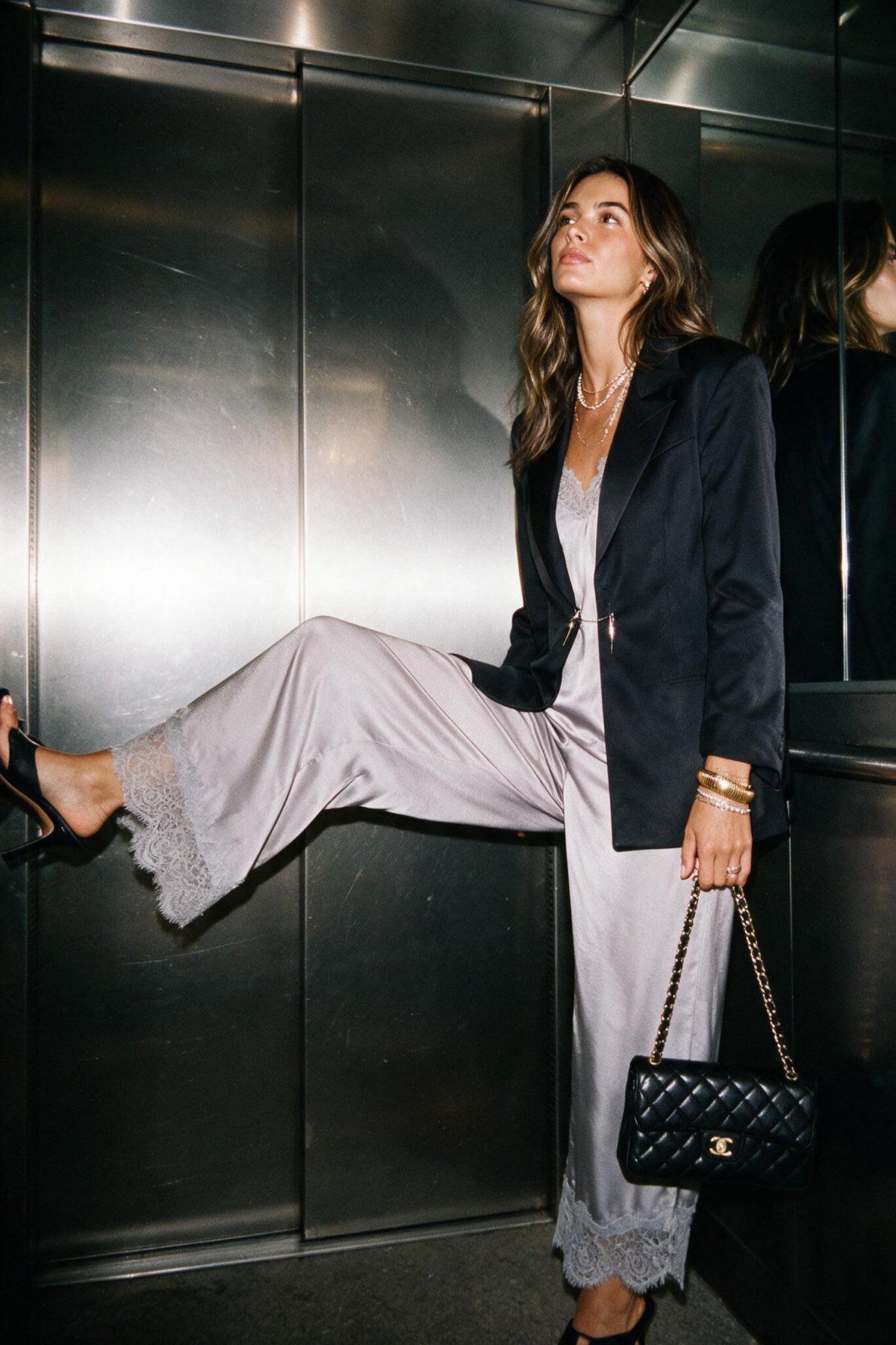 A woman posing in a metallic elevator with her leg extended, wearing a black blazer, light gray lace-trimmed pants, and holding a black quilted Chanel shoulder bag.
