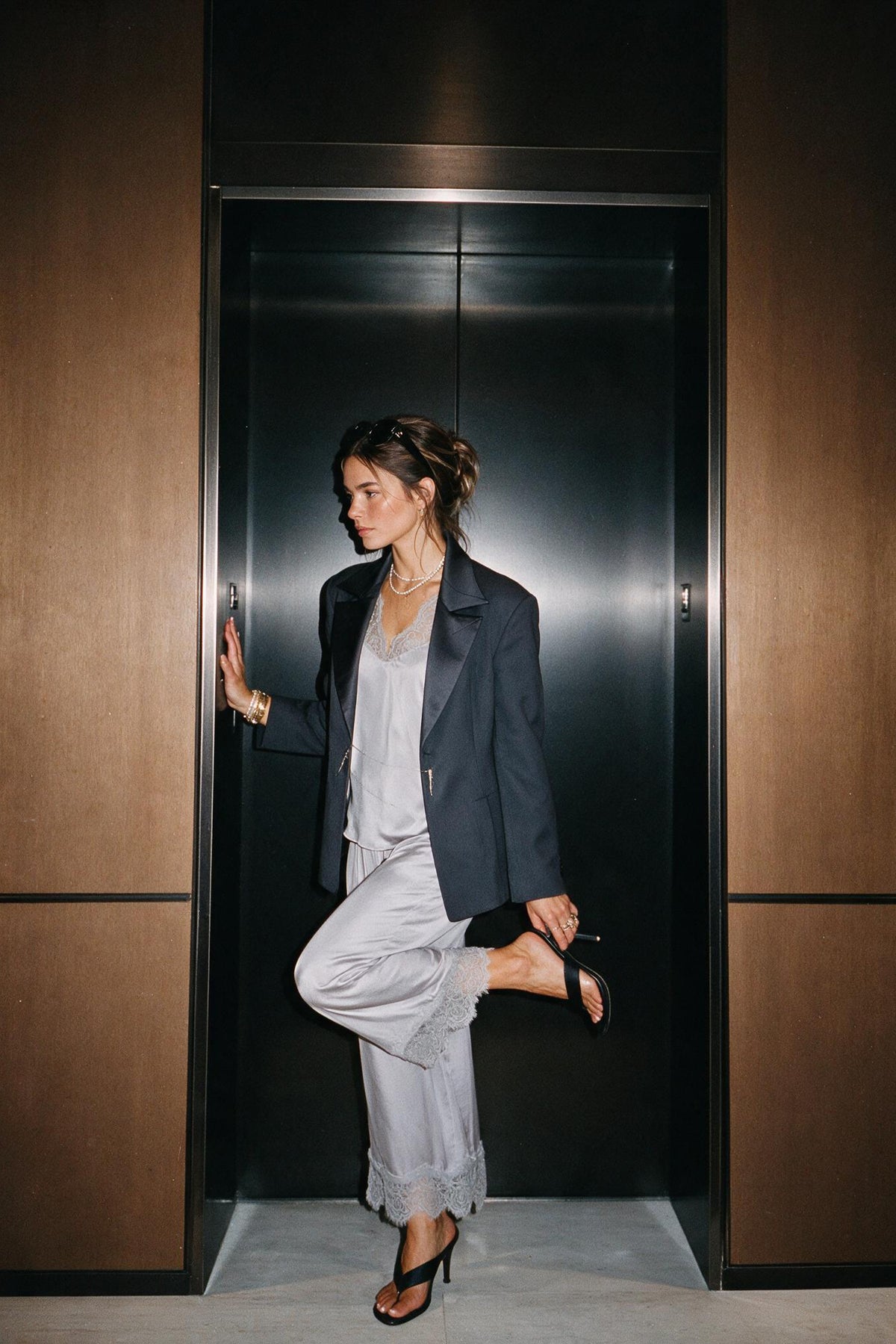 A woman posing in front of a dark metallic elevator door, wearing a dark navy blazer, a light gray top and matching wide-leg trousers, and black high-heeled sandals.
