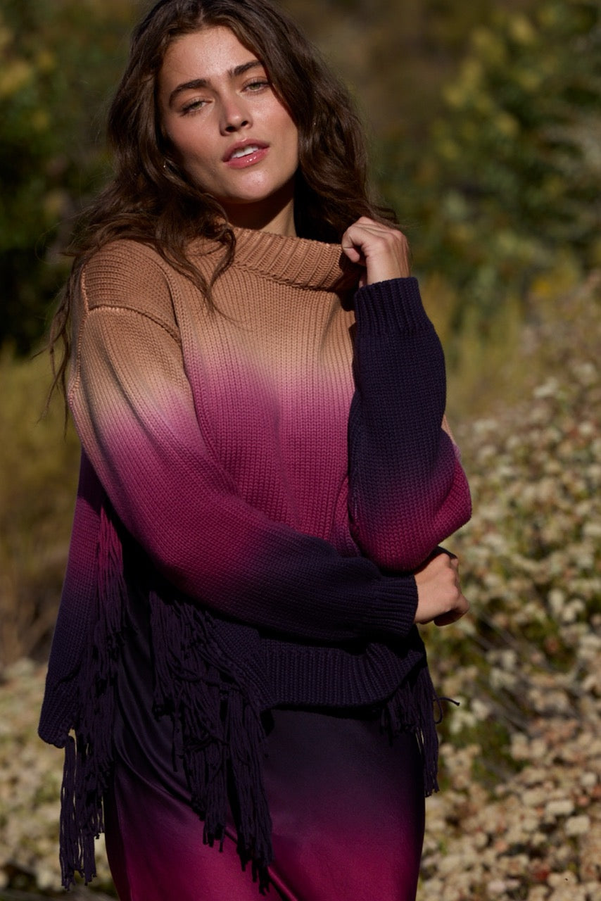 A woman wearing a ribbed knit sweater with a brown-to-purple-to-black ombre gradient and fringe trim, photographed outdoors against a blurred natural background.