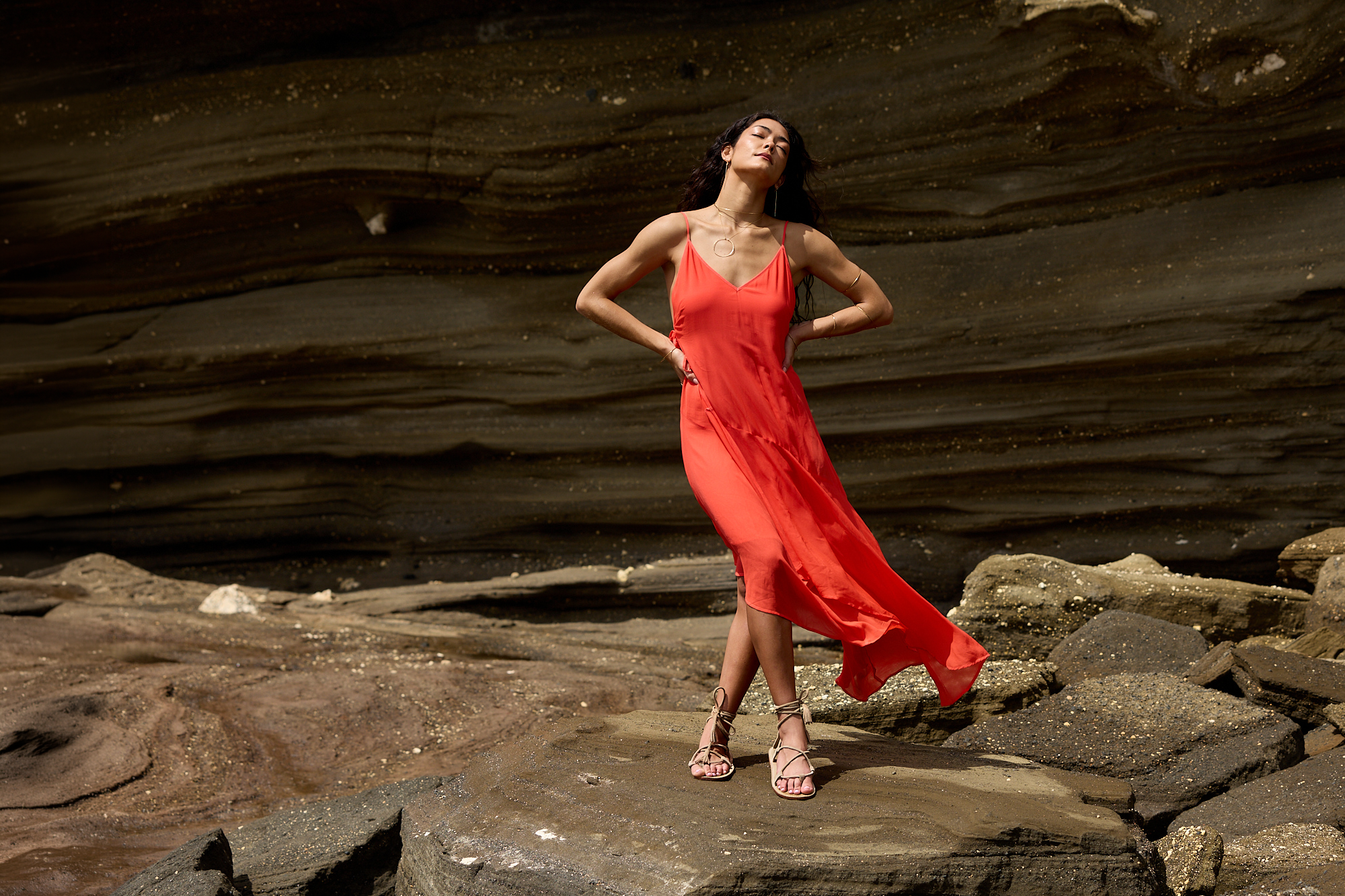 ADA compliant: Shop All collection. Woman wearing a chiffon flowy deep orange dress standing on boulders near the ocean.