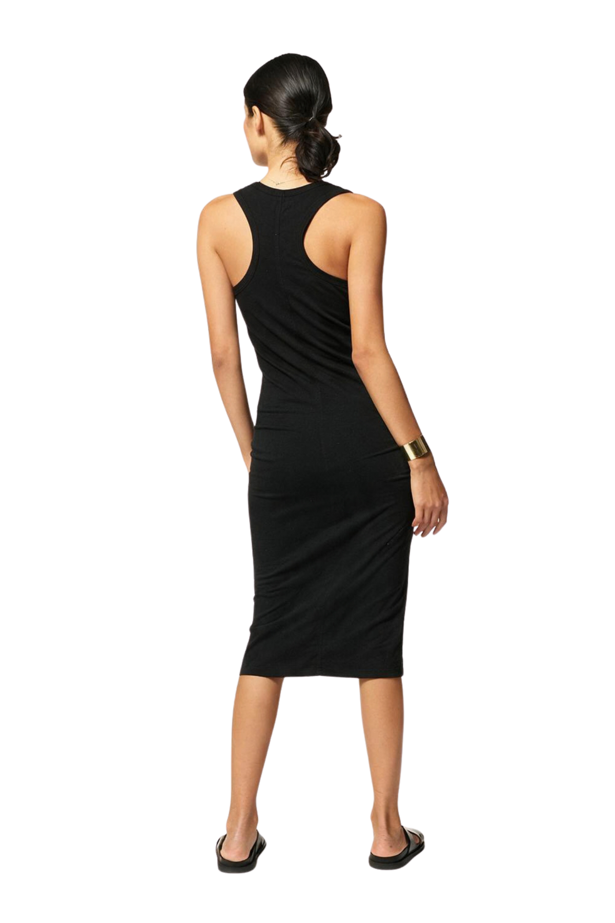 A woman viewed from the back wearing a sleeveless black midi dress with a wide shoulder cutout and a curved back design, paired with black flat sandals and a gold bracelet, on a white background.