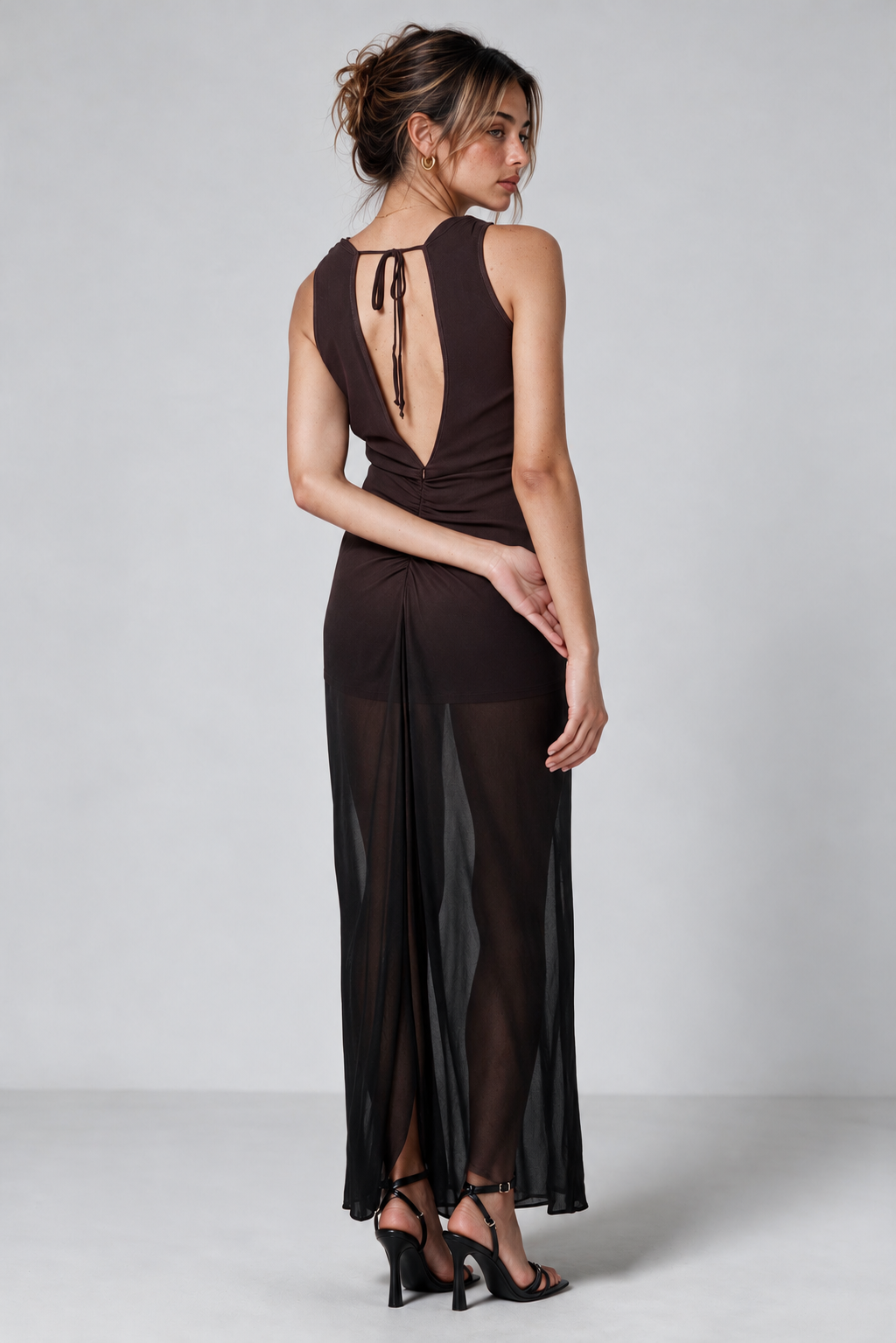 A woman modeling a dark brown ombre sleeveless maxi dress from the back, featuring a deep V-back with tie details and sheer mesh side panels, set against a light gray studio background.