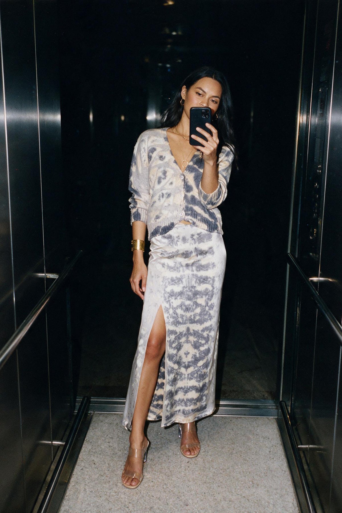 A woman takes a mirror selfie in an elevator wearing a long-sleeve cardigan and matching maxi skirt with a gray and white tie-dye pattern and a high side slit.