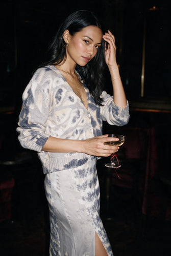 A woman wearing a light gray and navy blue tie-dye knit cardigan and matching skirt, holding a wine glass in a dimly lit indoor setting.