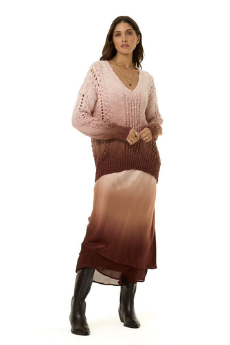 A woman modeling a long-sleeve knit sweater and matching midi skirt set in a dusty pink and brown ombre gradient, paired with black knee-high boots, against a plain white background.