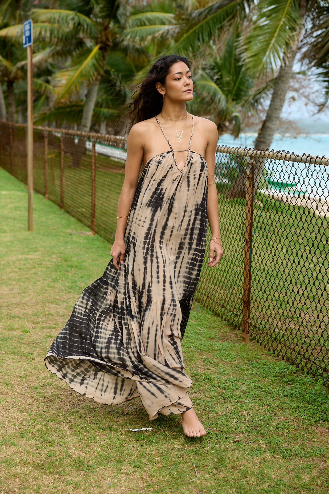 Woman wearing a chiffon flowy tan and black dress in a grassy scene with the ocean in the background