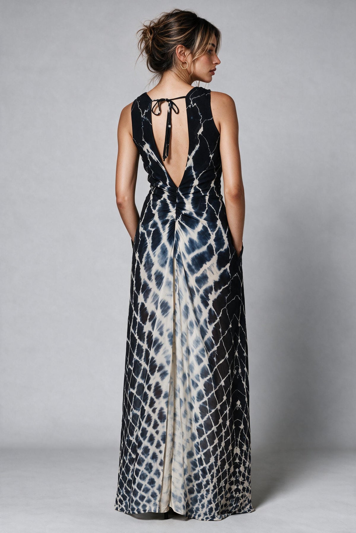 Backside of a woman modeling a sleeveless maxi dress with a black and off-white tie-dye pattern, standing against a neutral gray studio background.