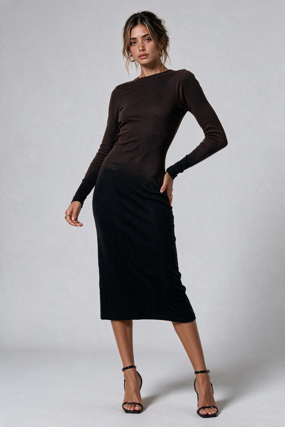A woman modeling a long-sleeve midi dress with a dark brown ombre effect fading into black at the skirt, paired with black strappy heels against a light gray studio background.