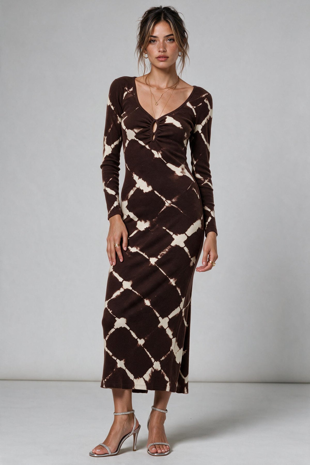 A woman modeling a long-sleeve midi dress with a brown and beige tie-dye diamond pattern, standing against a light gray studio background.