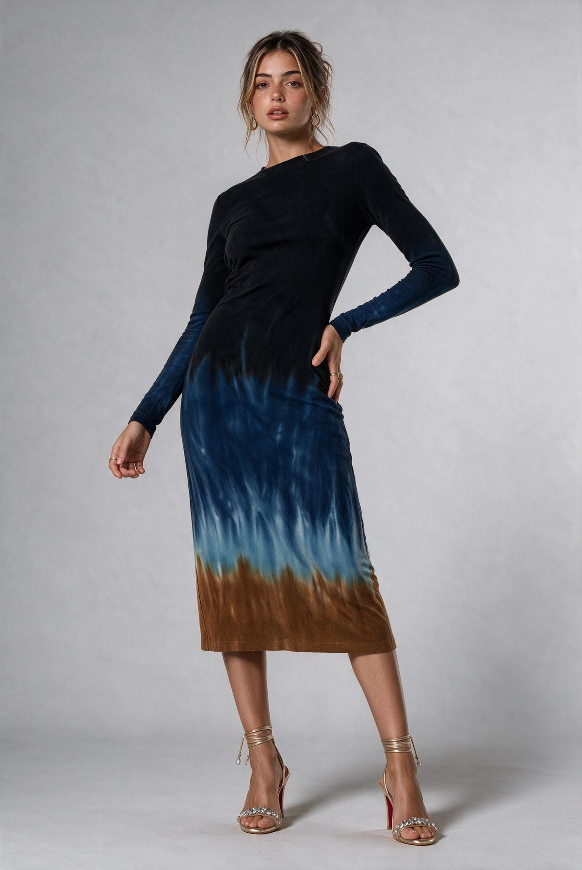 A woman modeling a long-sleeve midi dress with a black upper bodice and a blue-to-brown tie-dye skirt, paired with metallic high-heeled sandals against a light gray studio background.