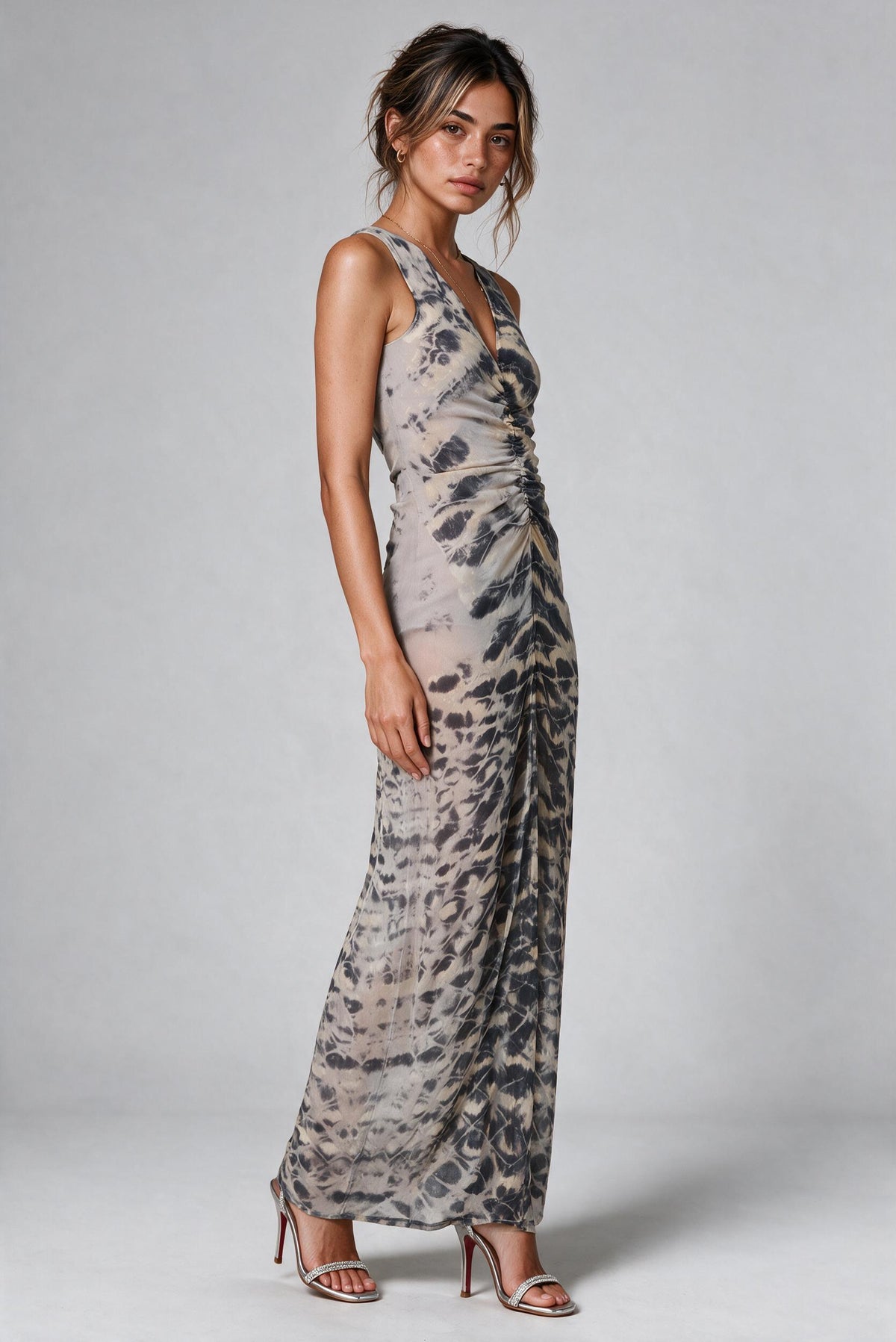 A woman modeling a sleeveless, floor-length dress with a gray and beige tie-dye pattern, paired with metallic silver high-heeled sandals, against a light gray studio background.