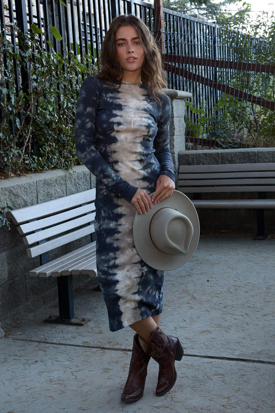 A woman stands outdoors in a park-like setting wearing a long-sleeve midi dress with a blue and beige tie-dye pattern, holding a wide-brimmed beige hat and wearing brown cowboy boots.