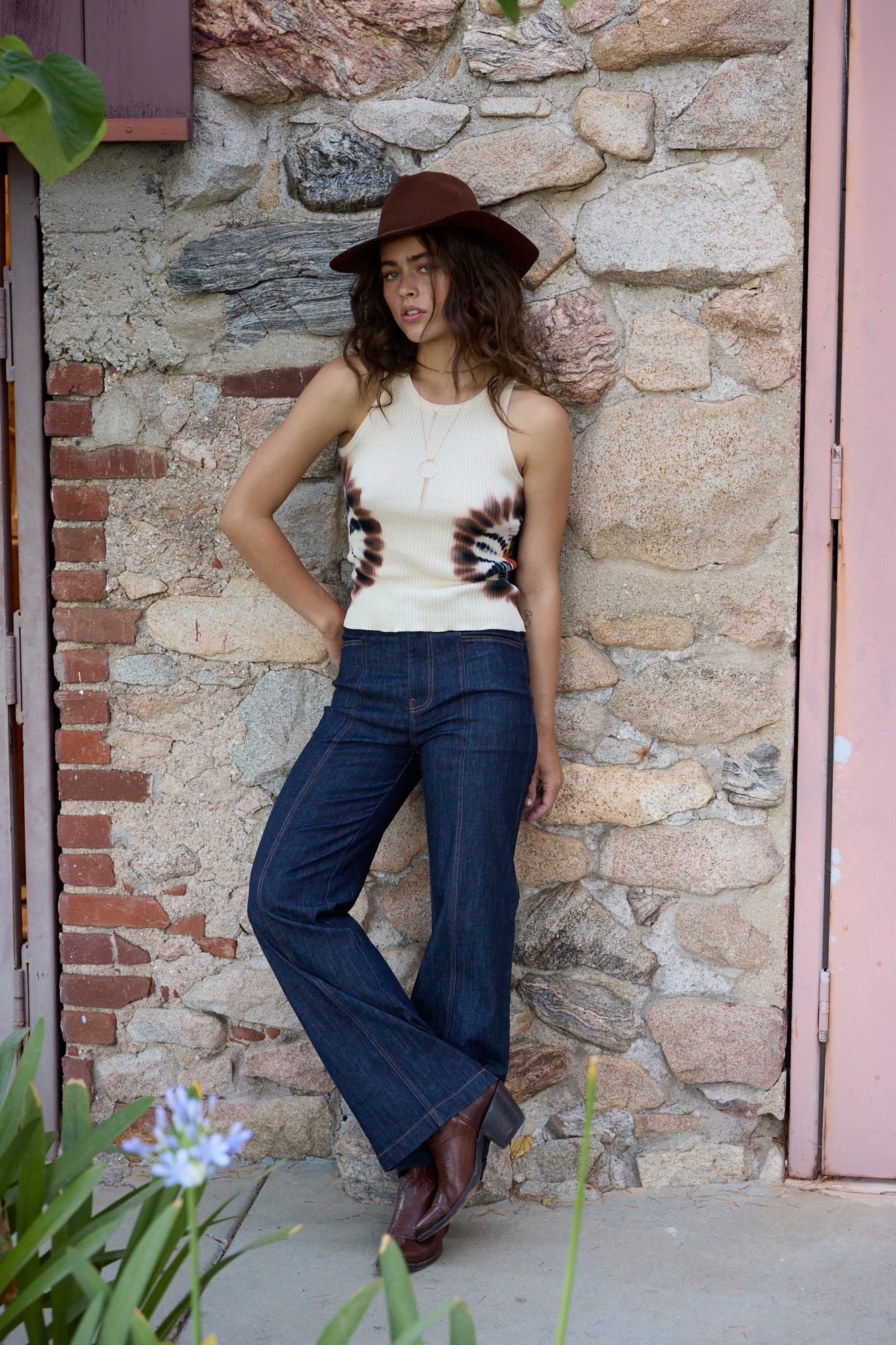 A woman stands against a rustic stone wall wearing a cream sleeveless top with a brown and black spiral print, dark blue wide-leg jeans, and a brown hat.