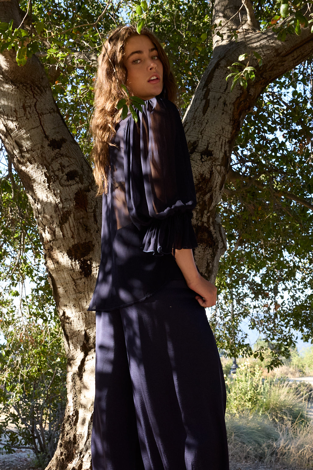 A woman wearing a dark navy blue long-sleeve blouse with a sheer ruffled collar and matching dark skirt, standing outdoors in front of a large tree with dappled sunlight.