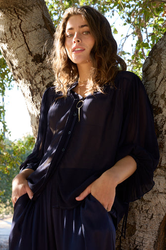 A woman wearing a dark navy blue sheer button-up shirt and matching skirt, leaning against a tree trunk outdoors.