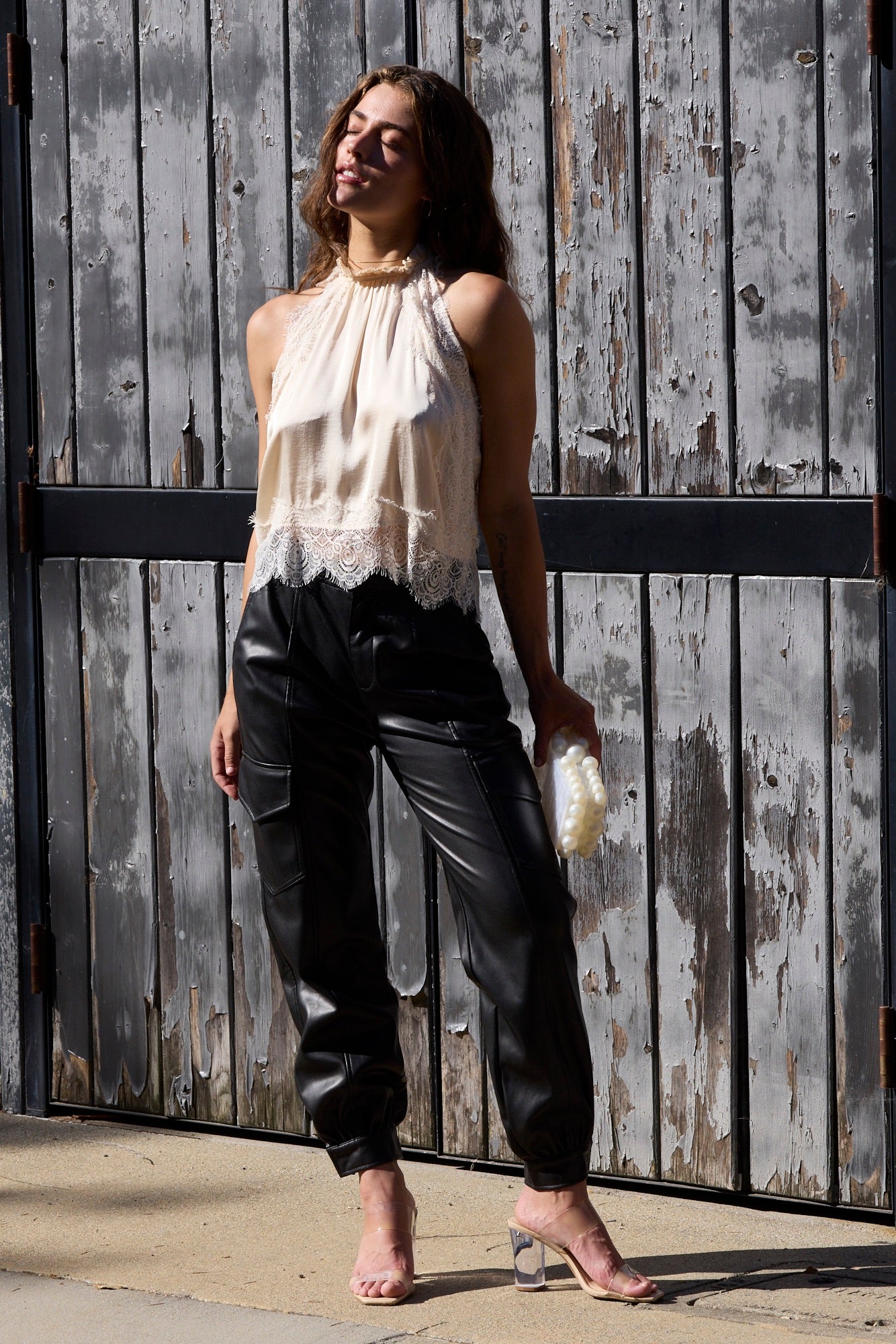 A woman stands outdoors in front of a weathered gray wooden fence, wearing a cream sleeveless lace-trim halter top, black leather pants, and clear high-heeled sandals.
