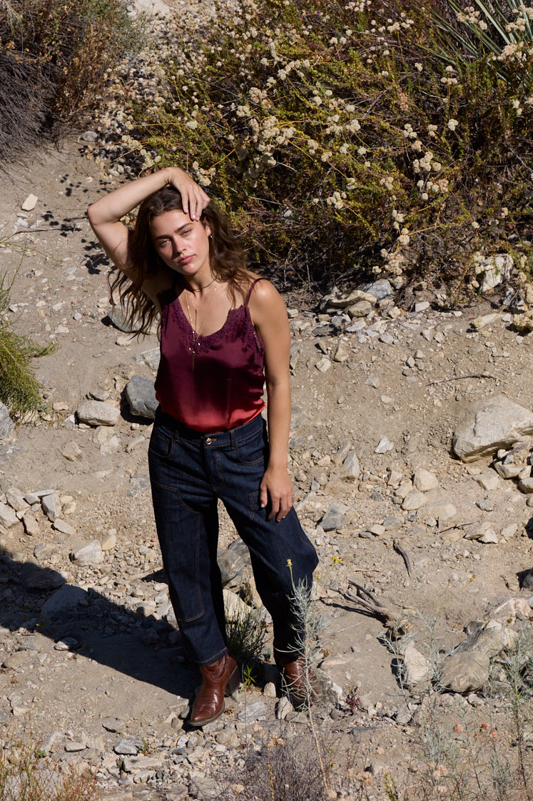 A woman posing outdoors on a rocky, sandy hillside wearing a dark burgundy satin tank top, dark blue jeans, and brown boots.