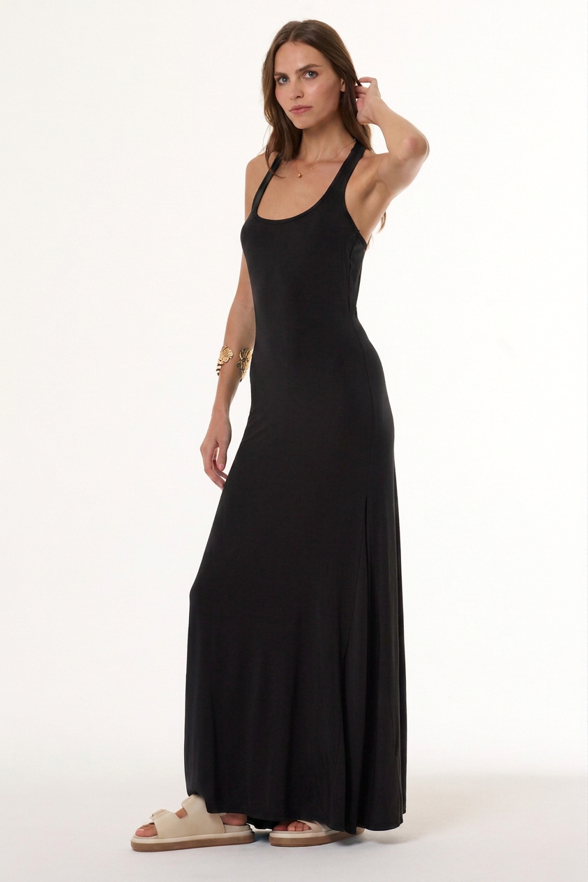 A woman modeling a sleeveless black maxi dress with a scoop neckline and a flowing skirt, paired with beige slide sandals, against a plain white studio background.