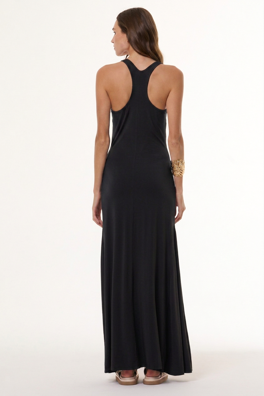 A woman viewed from the back wearing a sleeveless black maxi dress with a racerback cut and a high keyhole-style back, paired with beige flat sandals and a gold bracelet, against a plain white studio background.