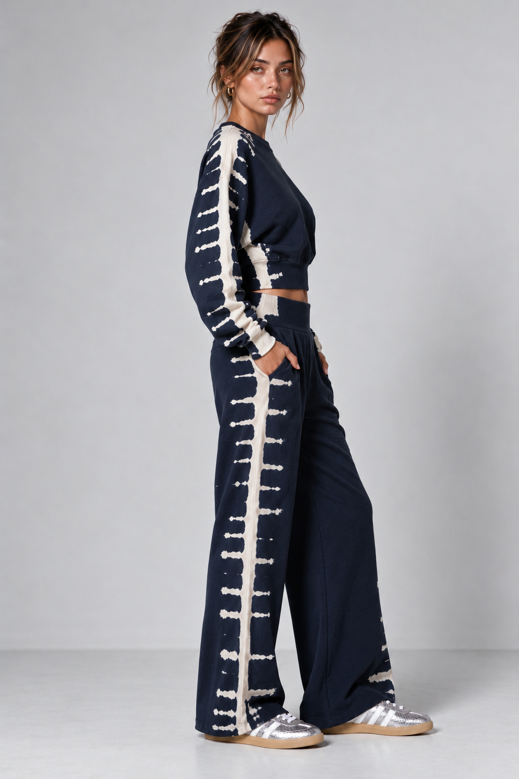 A woman modeling a navy blue two-piece set featuring a long-sleeve crop top and matching wide-leg pants, both detailed with vertical cream-colored embroidered stripes and decorative button-like motifs down the sides. She is wearing silver sneakers against a plain light gray studio background.