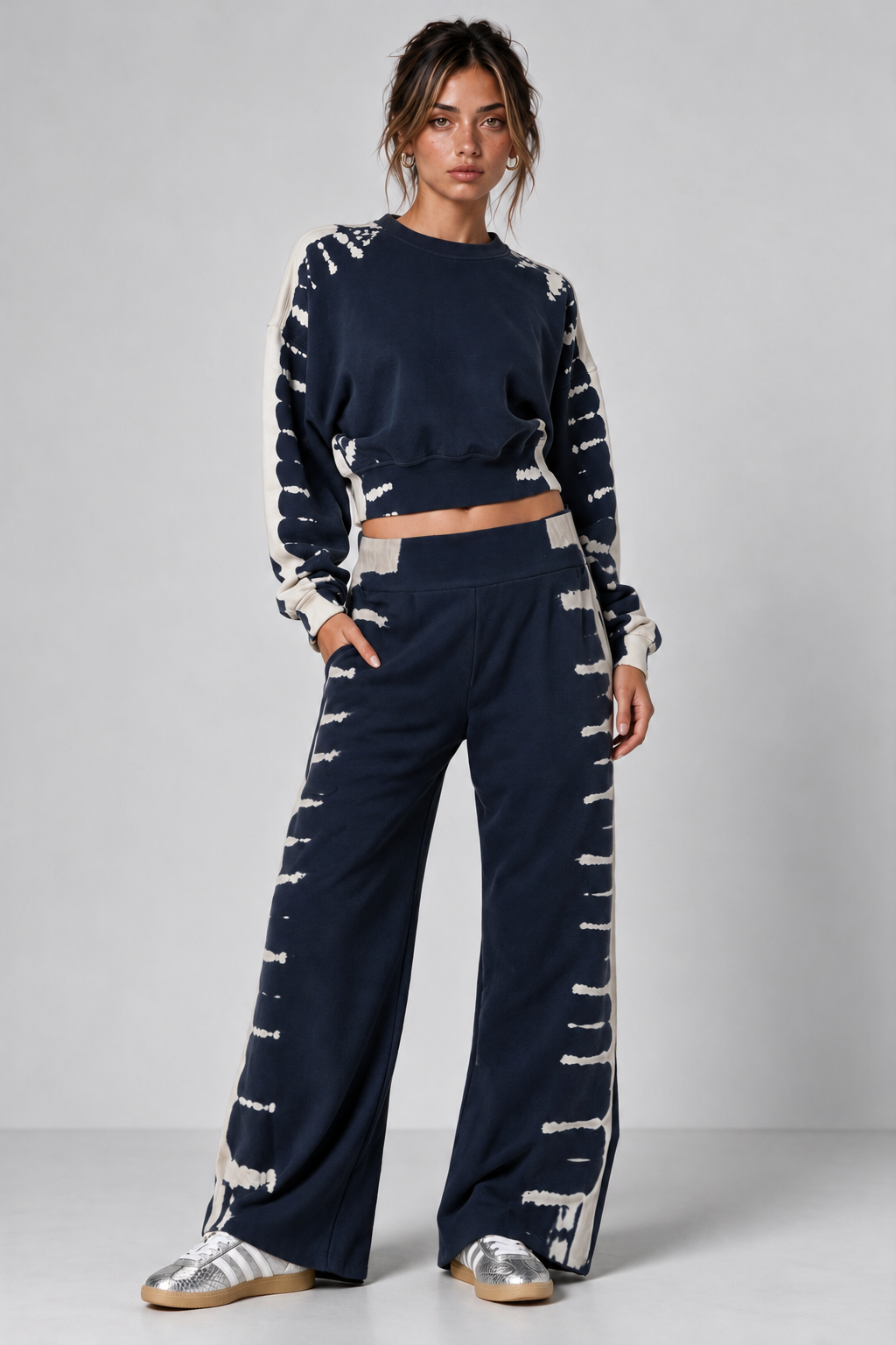 A model wearing a navy blue two-piece lounge set with distressed white tie-dye style patterns, consisting of a cropped long-sleeve sweatshirt and matching wide-leg pants, paired with silver sneakers against a light gray studio background.