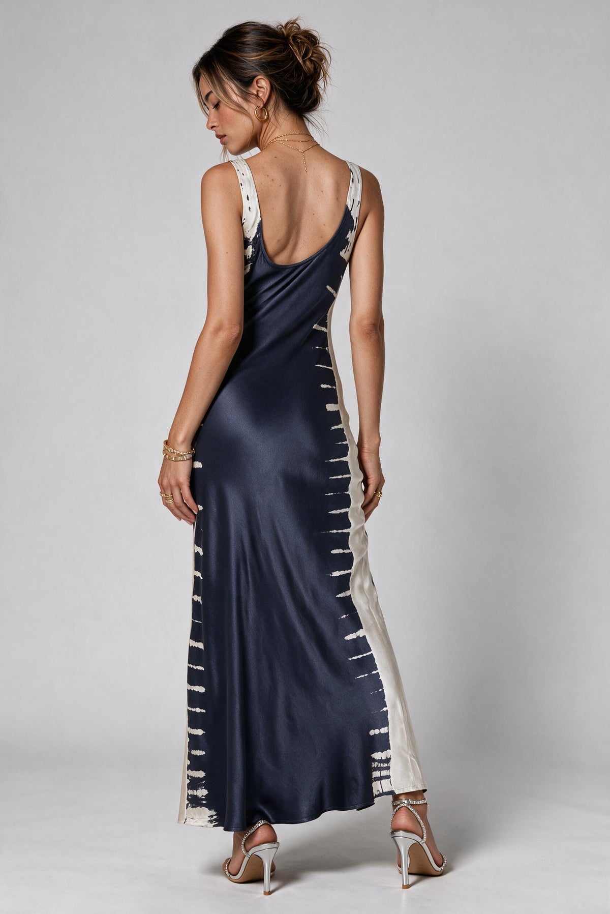 A woman modeling a navy blue satin maxi dress with off-white side panels and tie-dye style stripes, shown from the back against a light gray studio background.