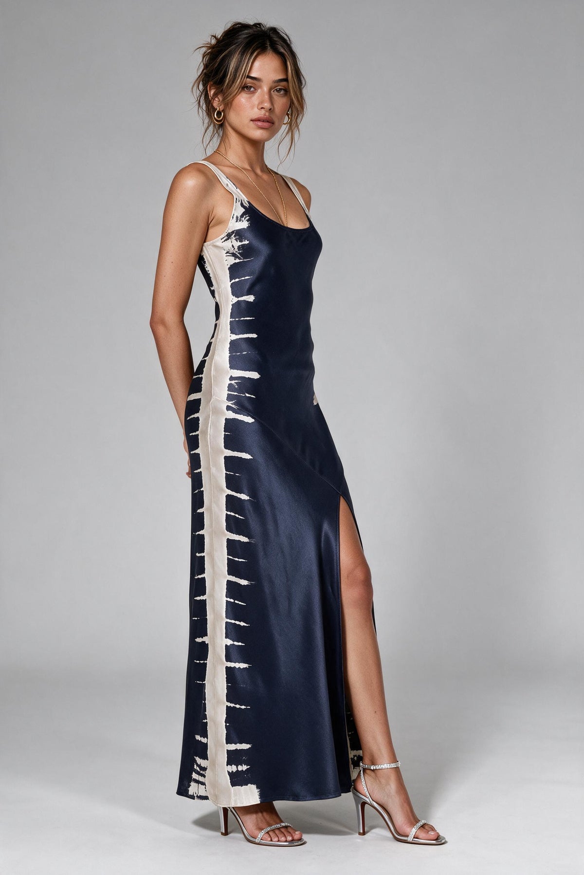 A woman models a navy blue satin slip dress with a high thigh slit and vertical white tie-dye style stripes down the sides, paired with clear high-heeled sandals against a neutral gray studio background.