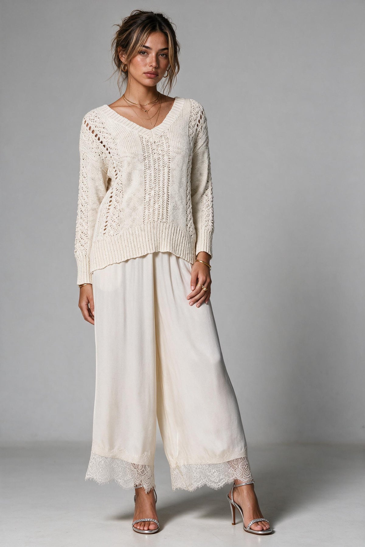 A woman modeling a cream-colored knit sweater and matching wide-leg pants with lace trim, paired with clear high-heeled sandals, against a neutral gray studio background.