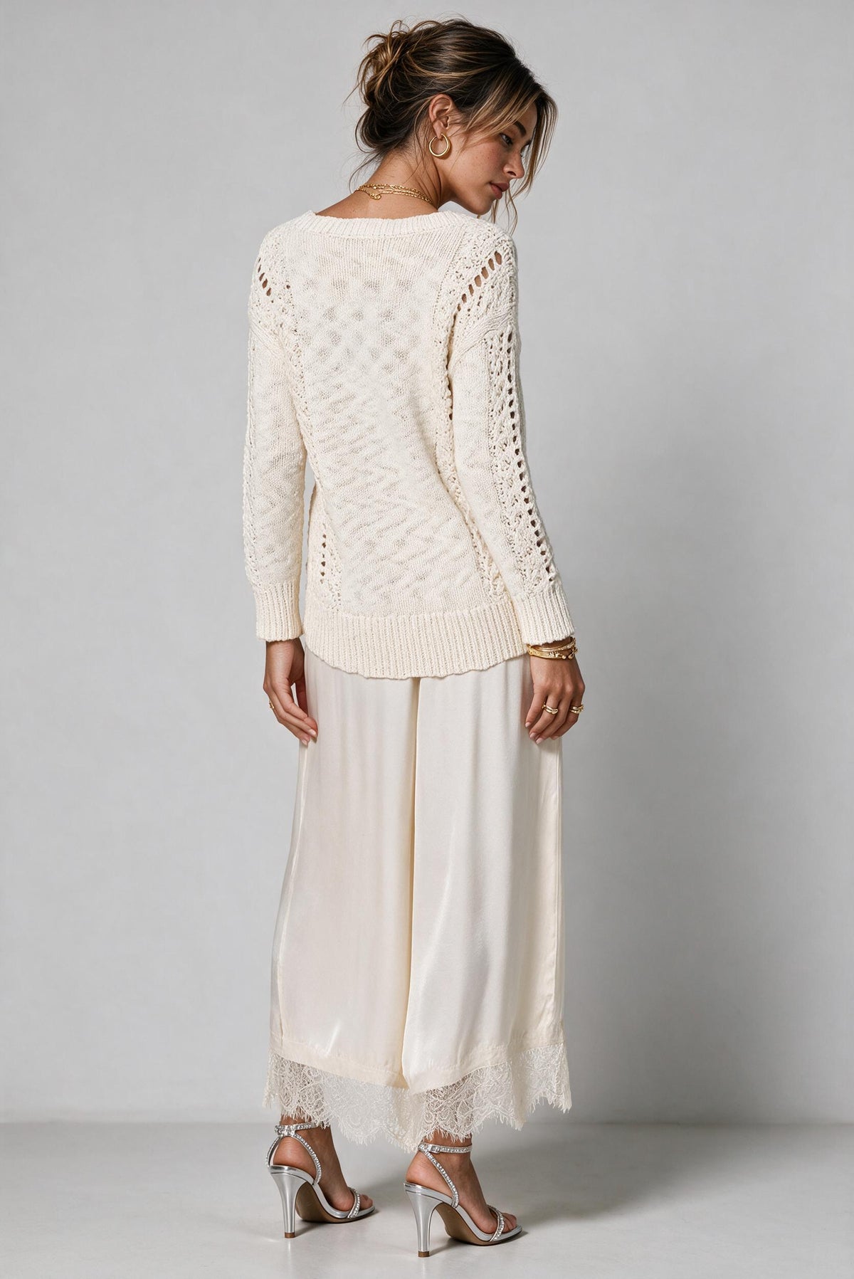 A woman shown from the back wearing a cream-colored knit sweater with lace-up cutouts, matching cream wide-leg pants with lace trim, and silver high-heeled sandals against a light gray studio background.
