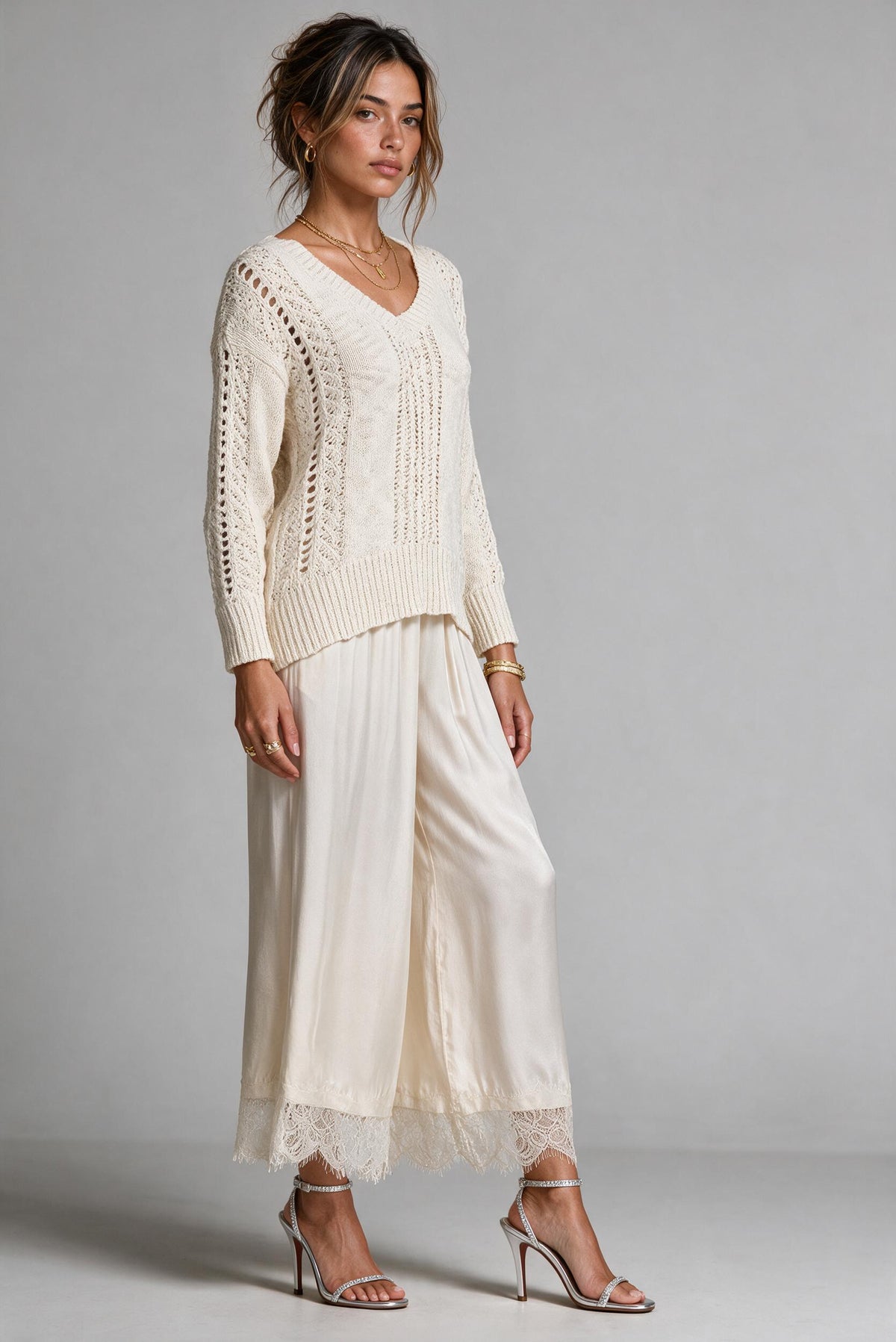 A woman modeling a cream-colored knit sweater and matching wide-leg trousers with lace trim, paired with silver high-heeled sandals, against a neutral gray studio background.
