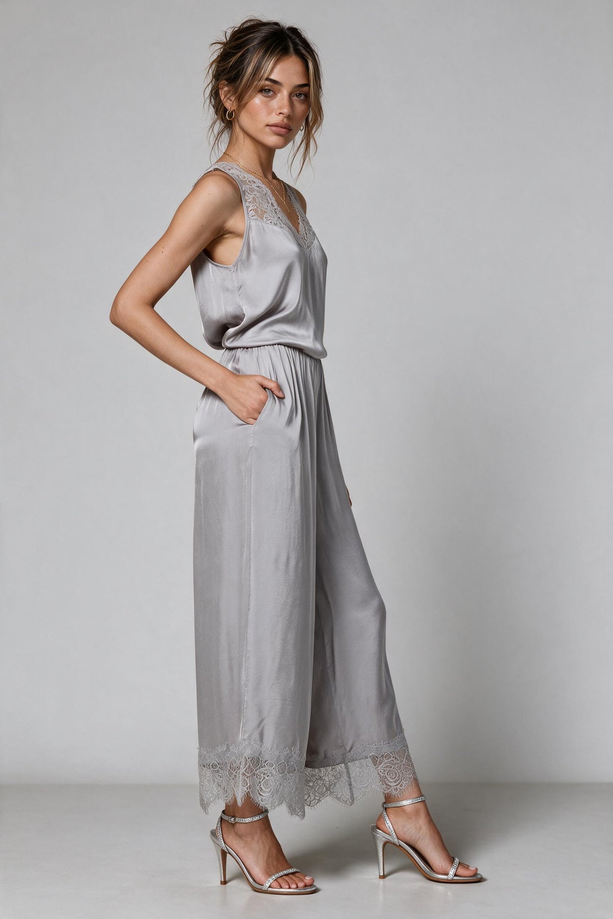 A woman modeling a light gray satin set with lace trim on the neckline and hem, paired with metallic heeled sandals, against a neutral gray studio background.