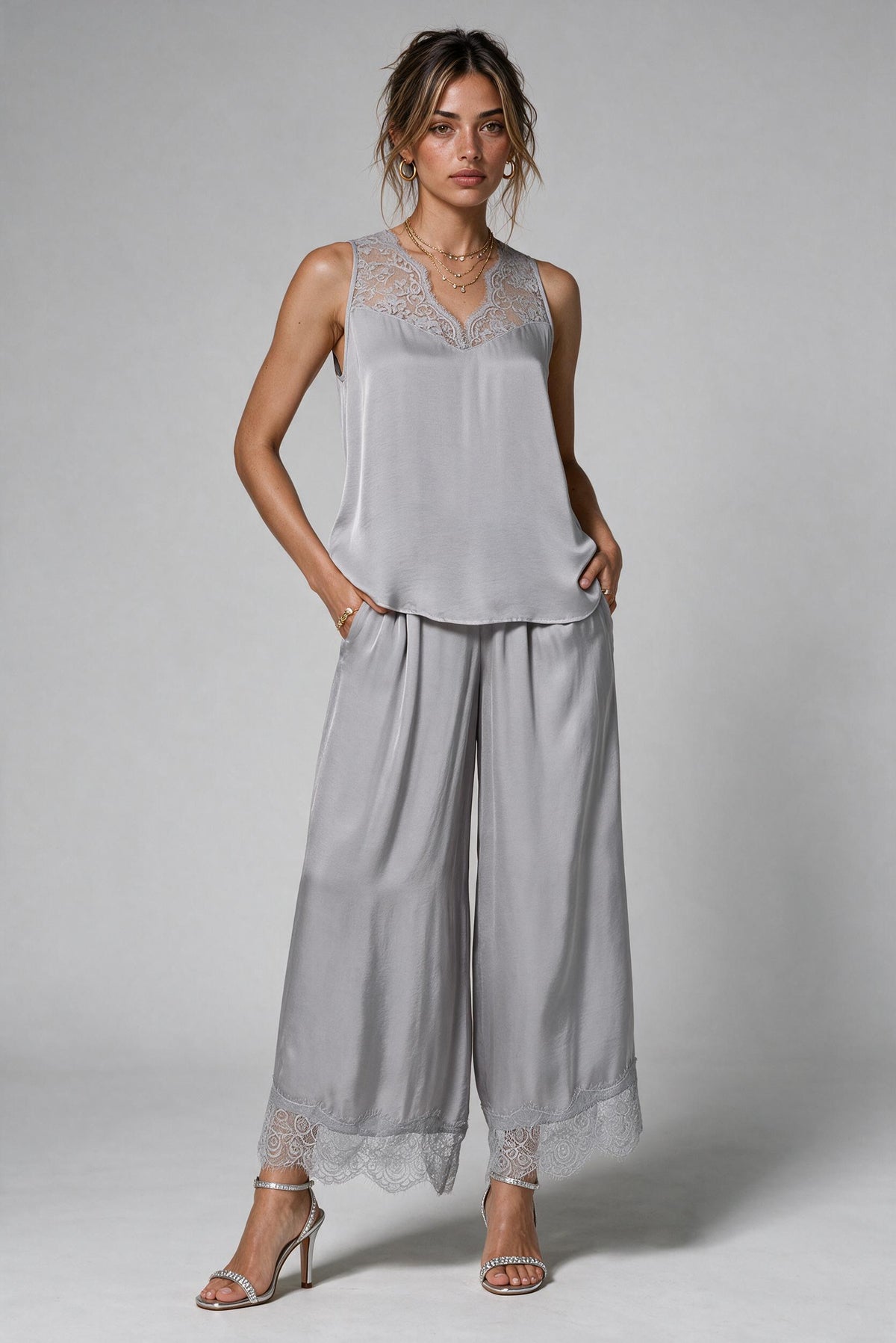 A woman modeling a light gray two-piece lounge set consisting of a sleeveless top with lace trim and matching wide-leg pants with lace hems, paired with silver high-heeled sandals against a neutral gray background.
