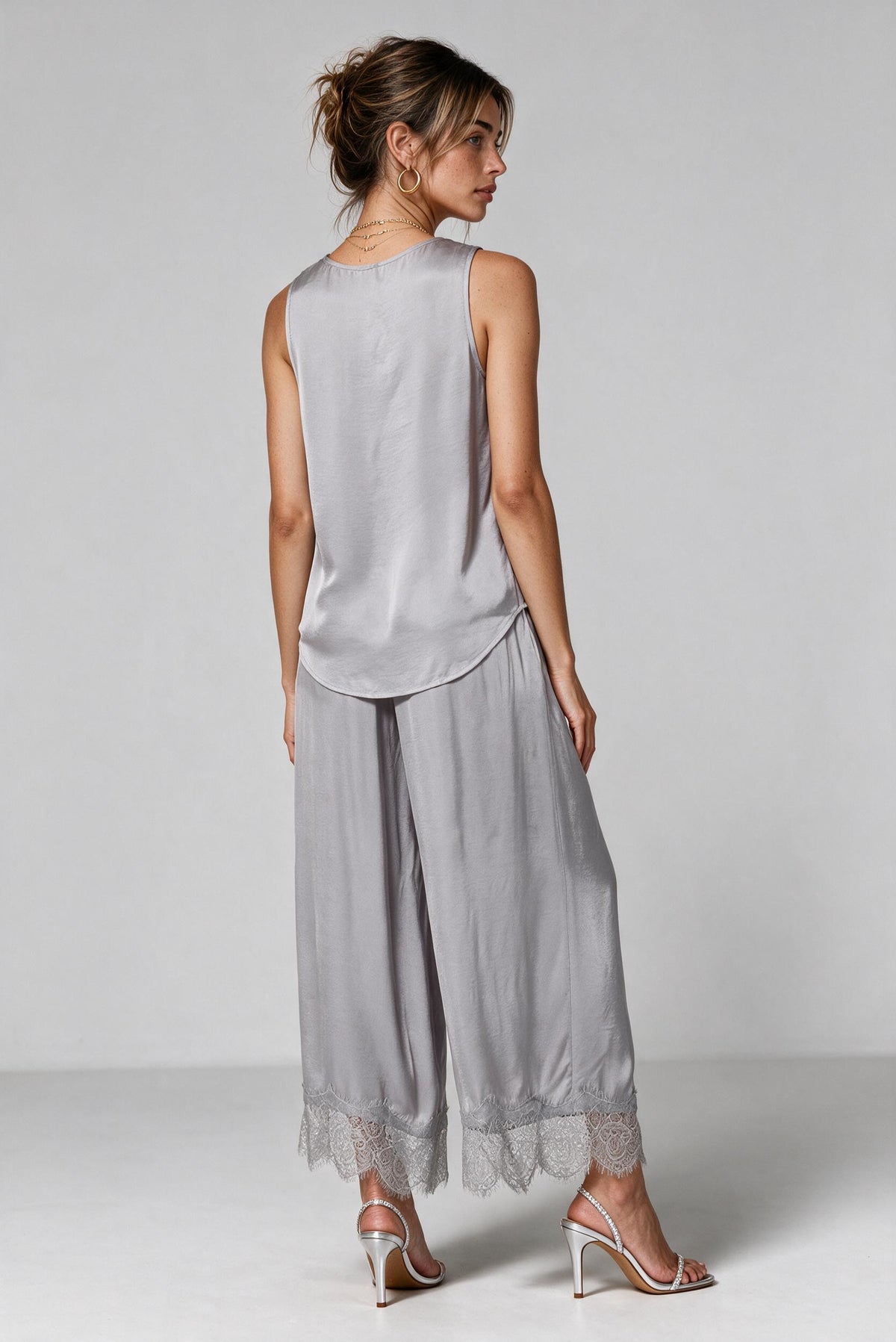 The backside os a woman modeling a light gray satin set with lace trim on the neckline and hem, paired with metallic heeled sandals, against a neutral gray studio background.