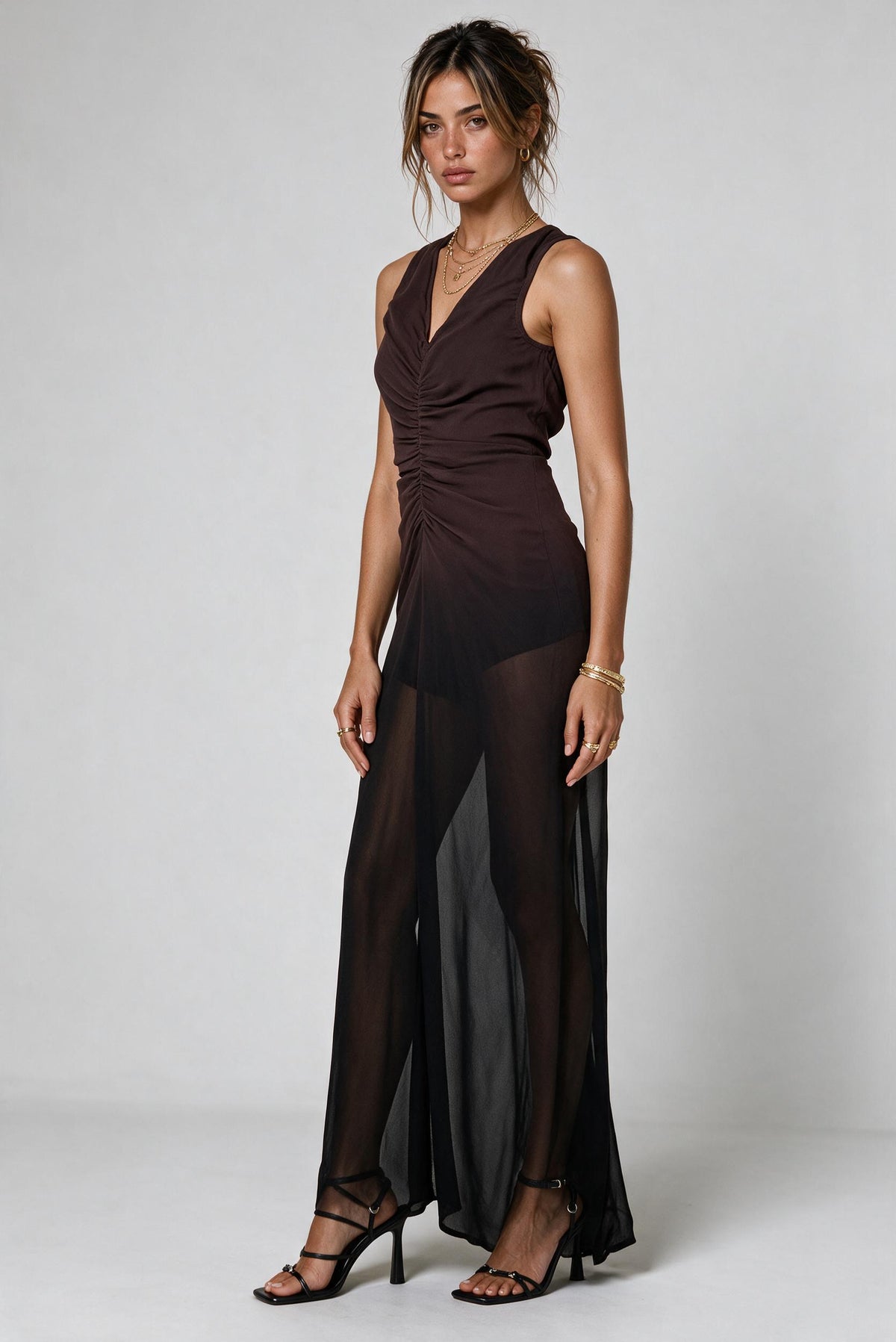 A woman modeling a sleeveless dark brown ombre maxi dress with a sheer black overlay skirt, photographed against a neutral light gray studio background.