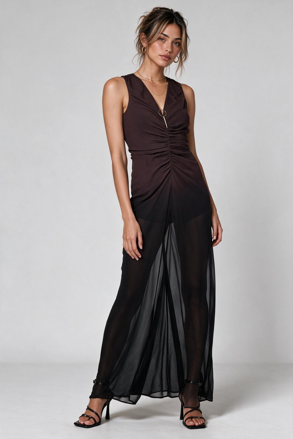 A woman modeling a sleeveless dark brown and black ombre draped maxi dress with sheer black overlay panels and black strappy heels, photographed against a light gray studio background.