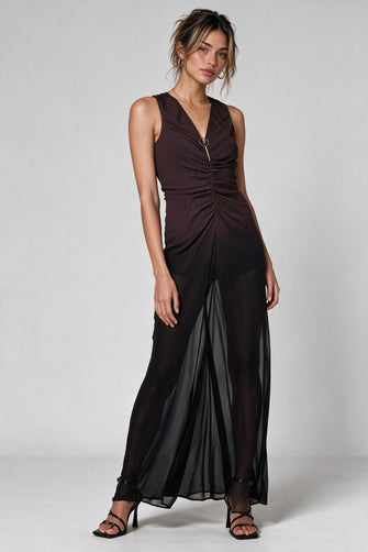 A woman modeling a sleeveless dark brown and black ombre draped maxi dress with sheer black overlay panels and black strappy heels, photographed against a light gray studio background.