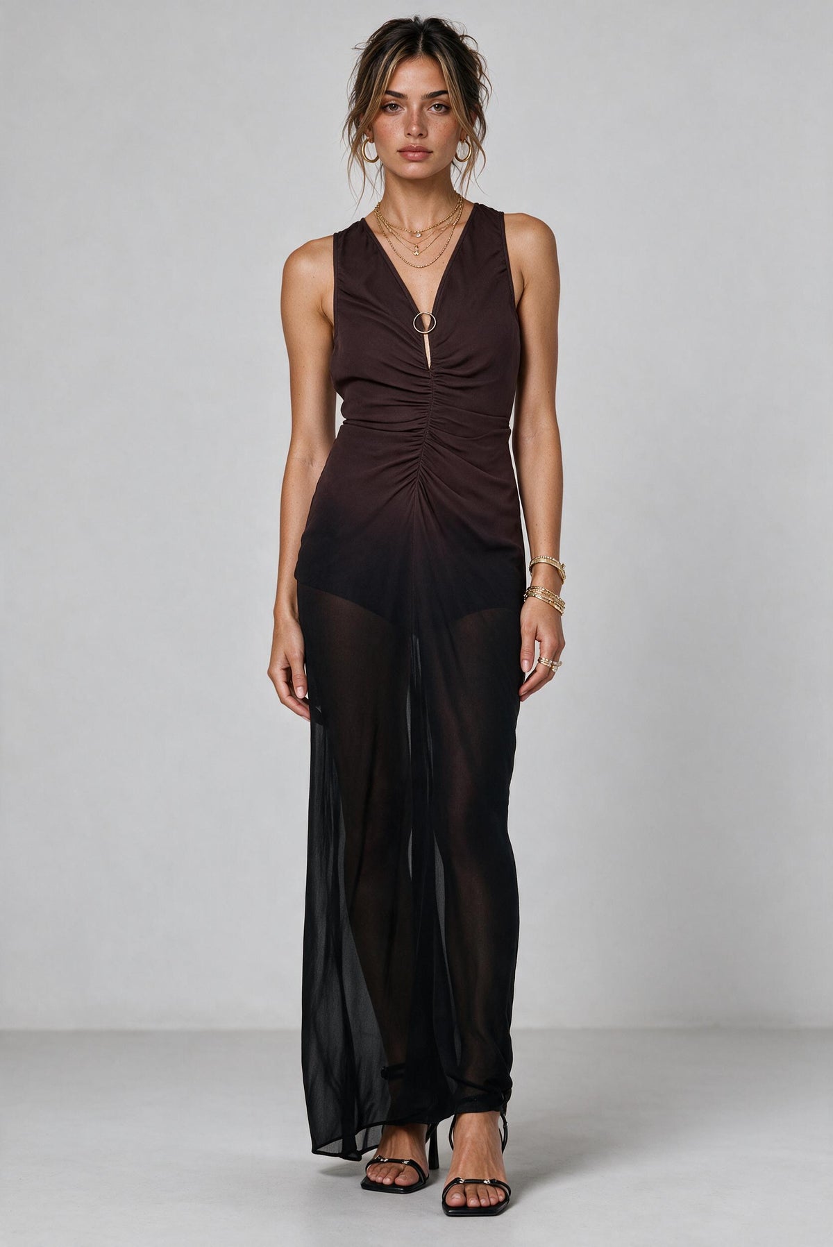 A woman models a sleeveless dark brown ombre ruched maxi dress with a sheer black overlay skirt, photographed against a neutral light gray studio background.