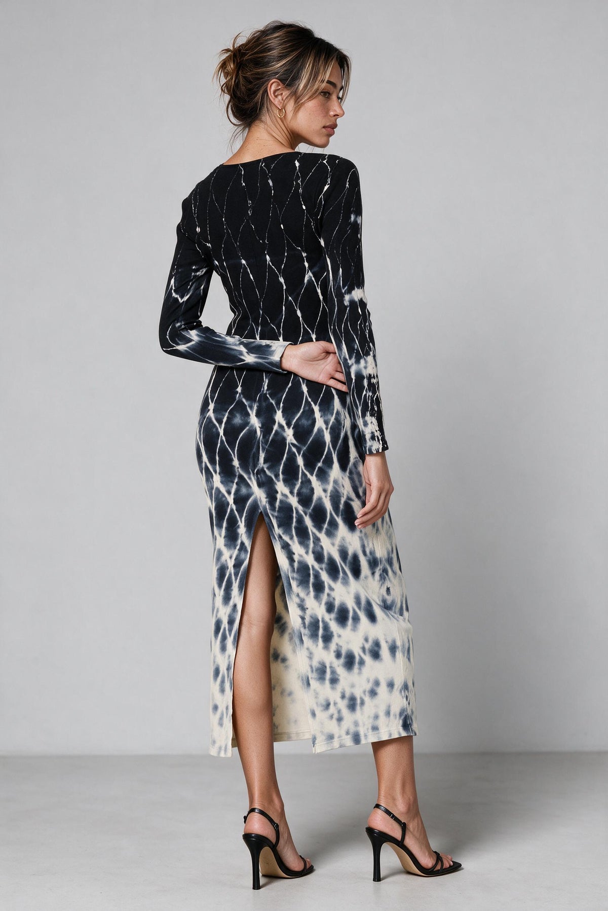 A woman modeling a long-sleeve midi dress with a black and off-white tie-dye pattern, shown from the back against a light gray studio background.