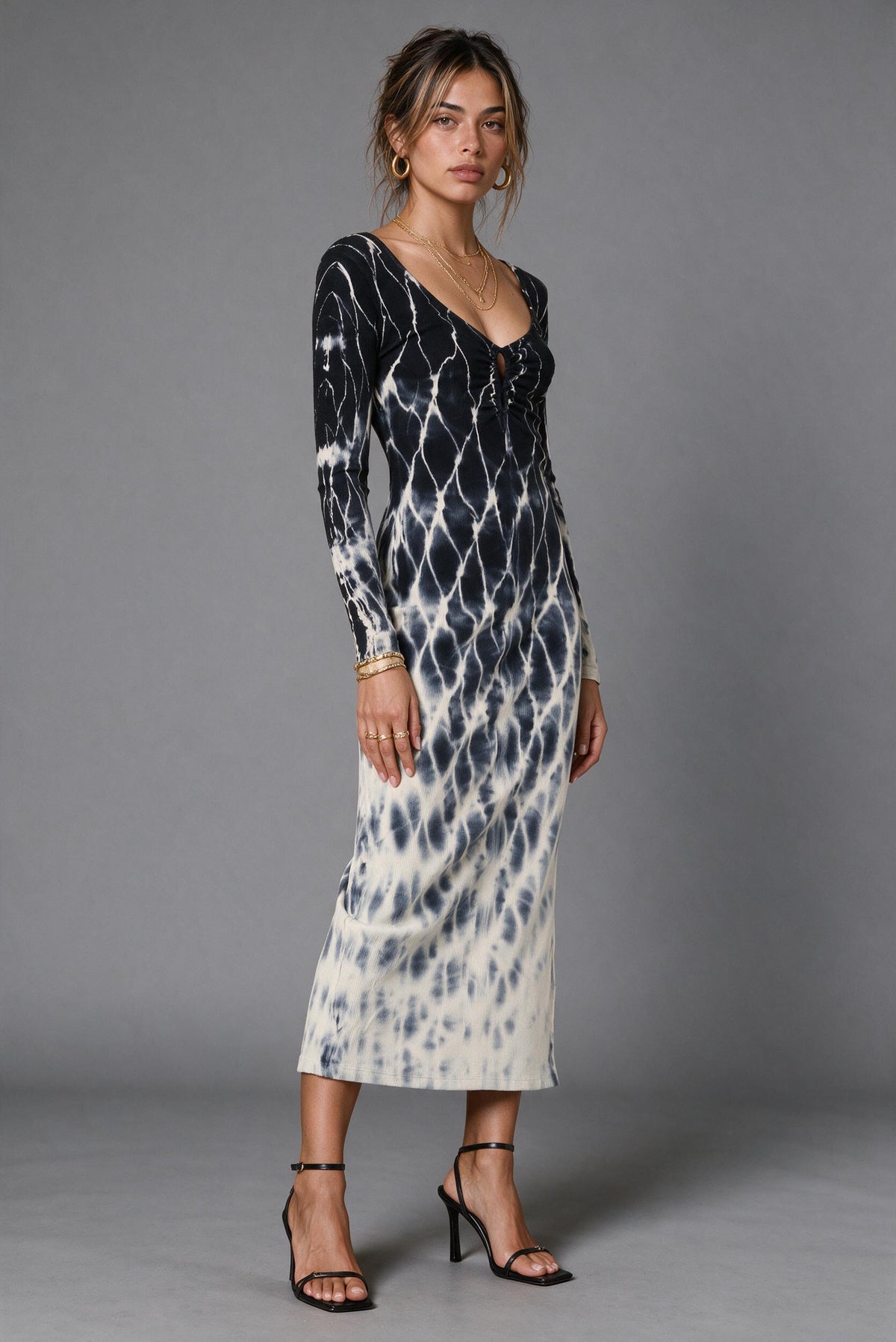A woman modeling a long-sleeve midi dress with a black and off-white tie-dye pattern, standing against a solid gray studio background.