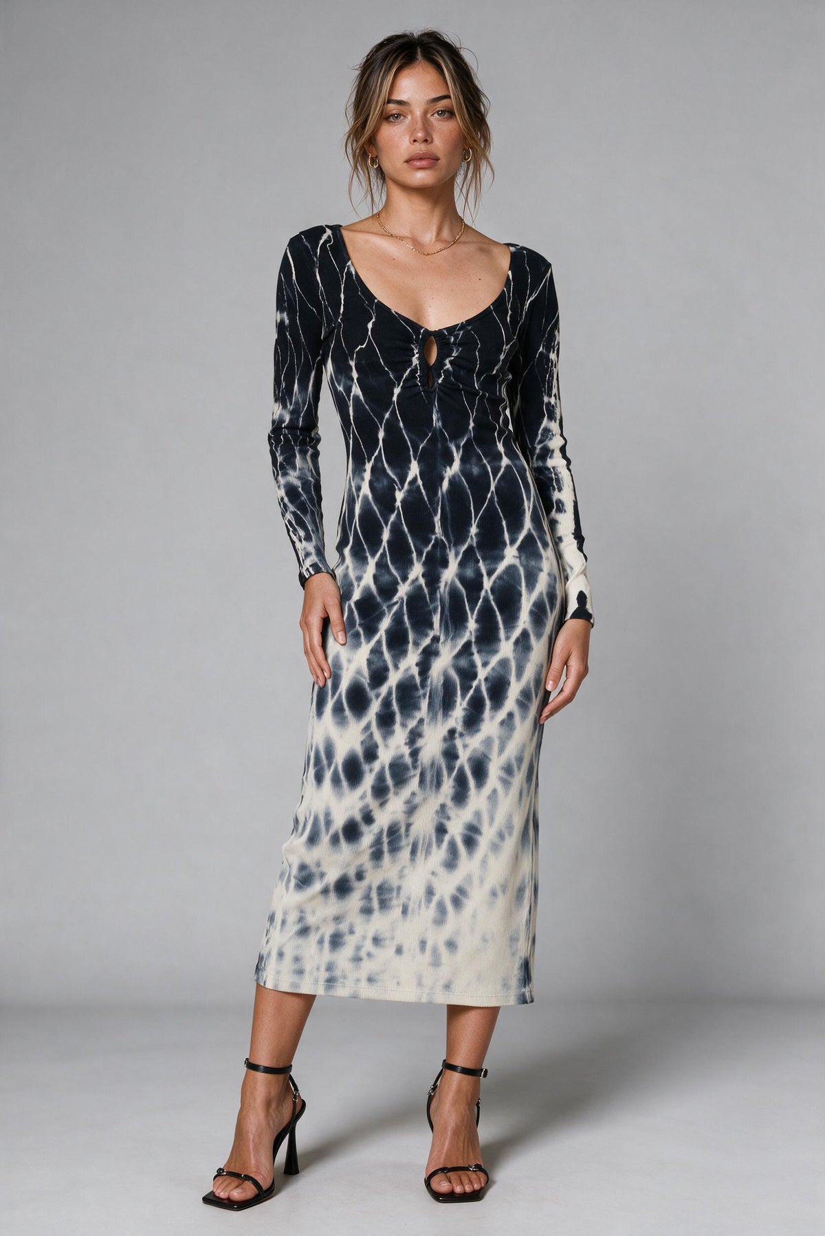 A woman modeling a long-sleeve midi dress with a black and off-white tie-dye pattern, standing against a solid gray studio background.