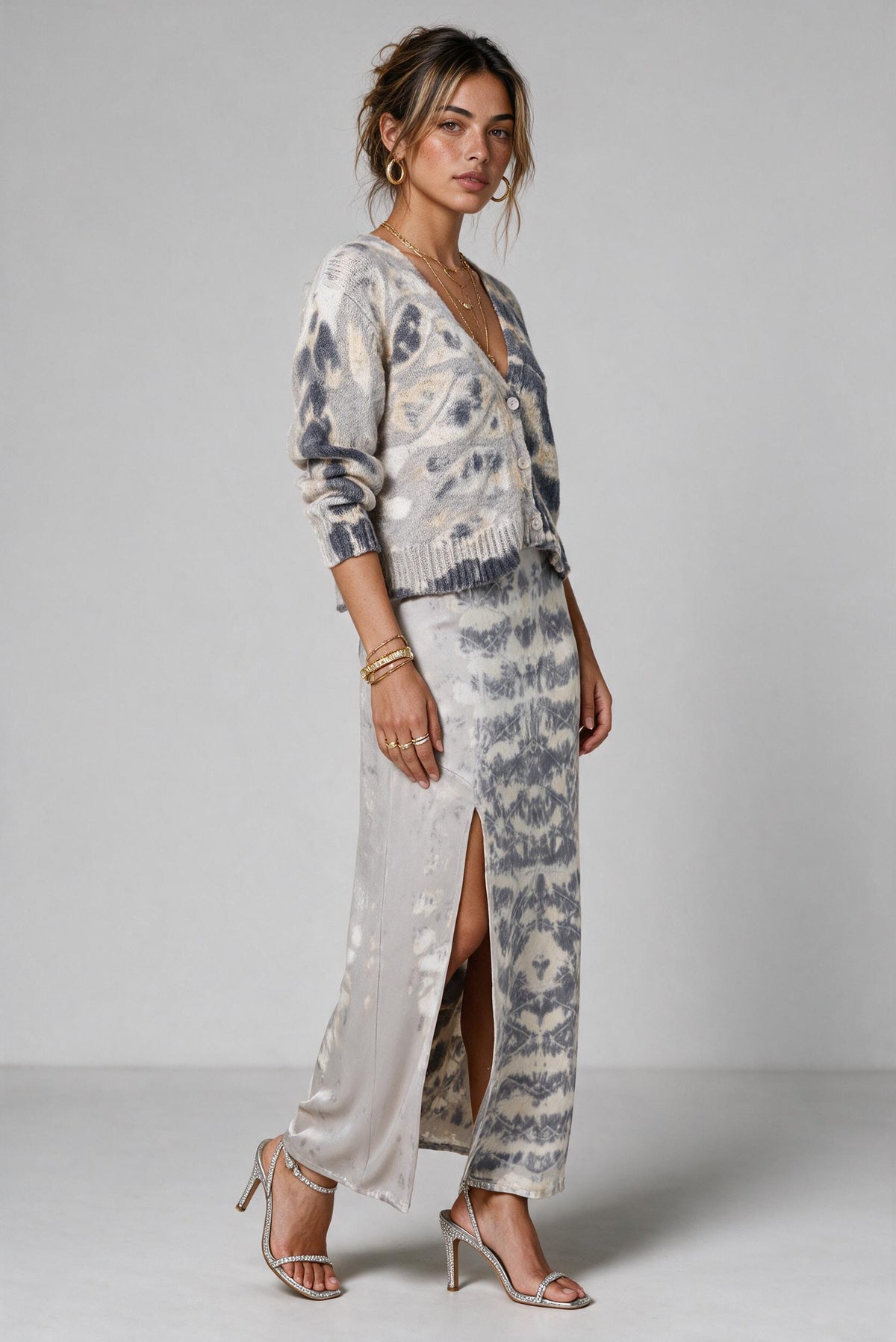 A woman models a matching two-piece knit and satin set featuring a gray and white abstract patterned cardigan and a matching high-slit skirt, styled with beige strappy heels against a neutral gray studio background.