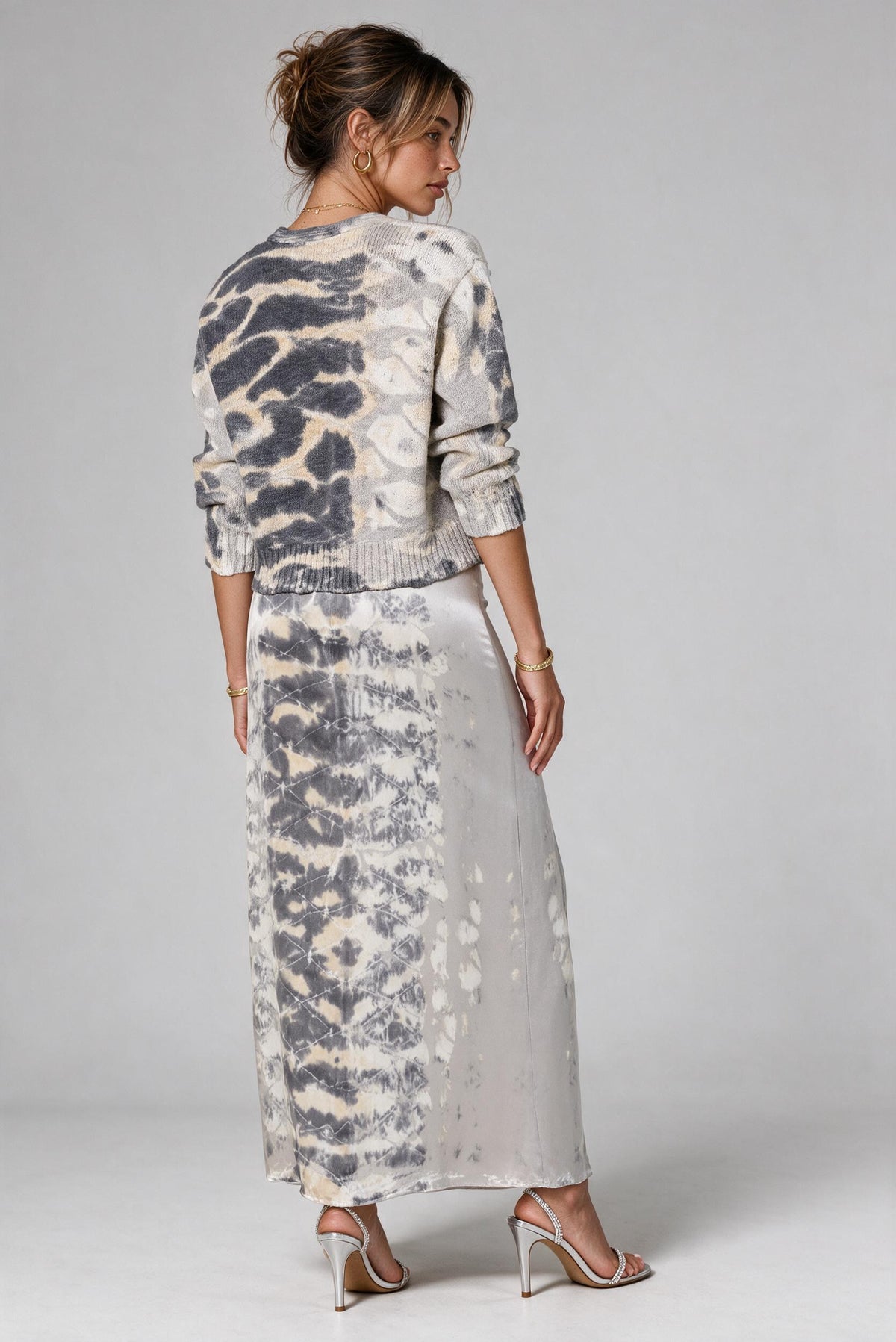 A woman modeling a matching two-piece knit set featuring a cropped long-sleeve cardigan and a maxi skirt, both in a gray, white, and beige animal print pattern, shown from a rear three-quarter angle against a light gray studio background.