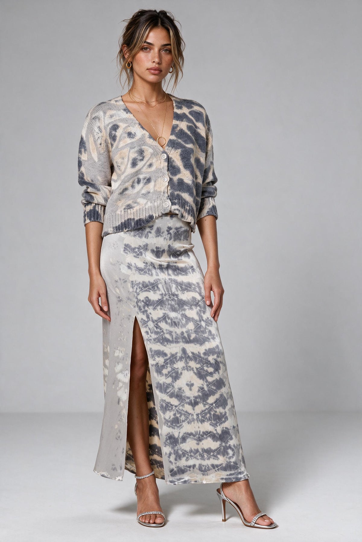 A woman models a matching two-piece knit set featuring a long-sleeve wrap cardigan and a high-slit maxi skirt, both in a gray and beige tie-dye pattern, set against a plain light gray studio background.