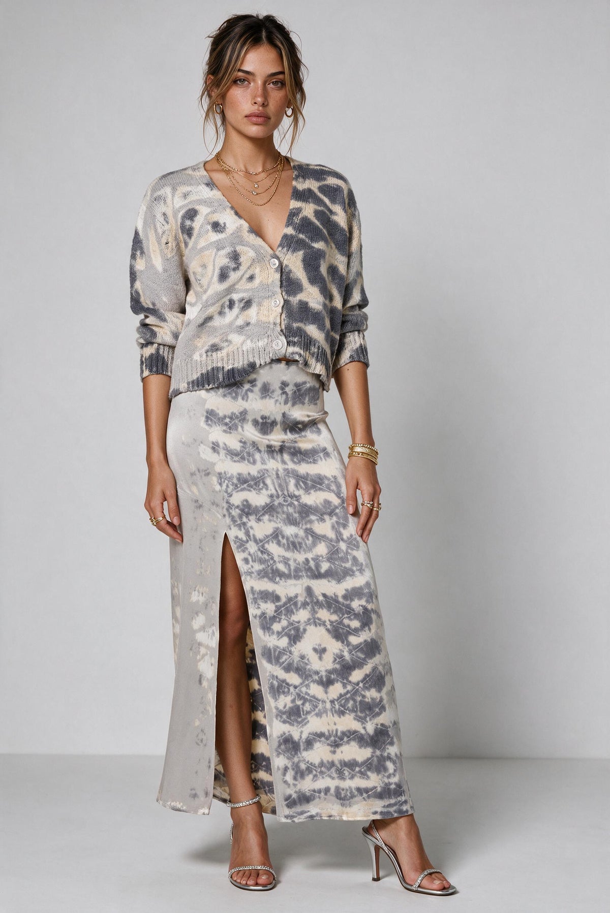 A woman models a matching two-piece knit set featuring a long-sleeve wrap cardigan and a high-slit maxi skirt, both in a gray and beige tie-dye pattern, set against a plain light gray studio background.