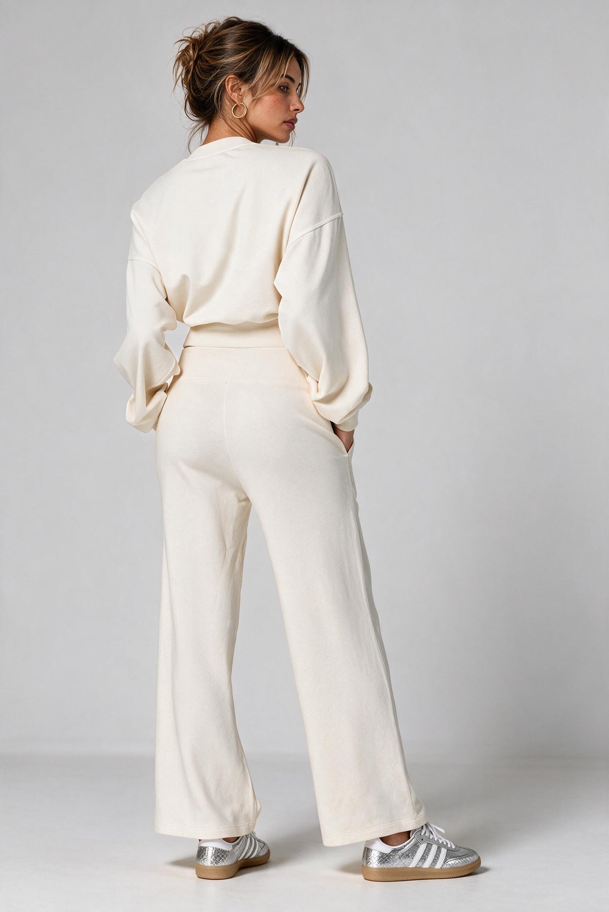 A woman shown from the back wearing a matching cream-colored lounge set consisting of a long-sleeve top and wide-leg trousers sweatpants, paired with silver sneakers, against a light gray studio background.
