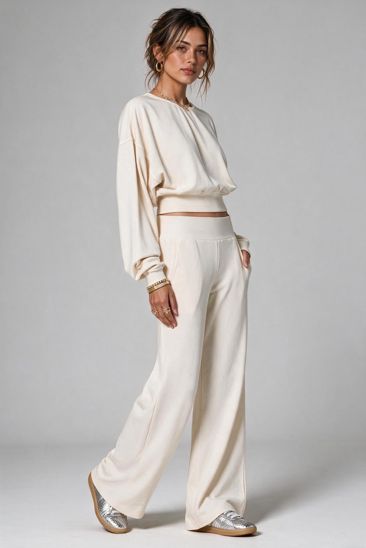 A woman modeling a cream-colored two-piece lounge set consisting of a long-sleeve cropped top and matching wide-leg pants, paired with metallic silver sneakers, against a neutral gray studio background.