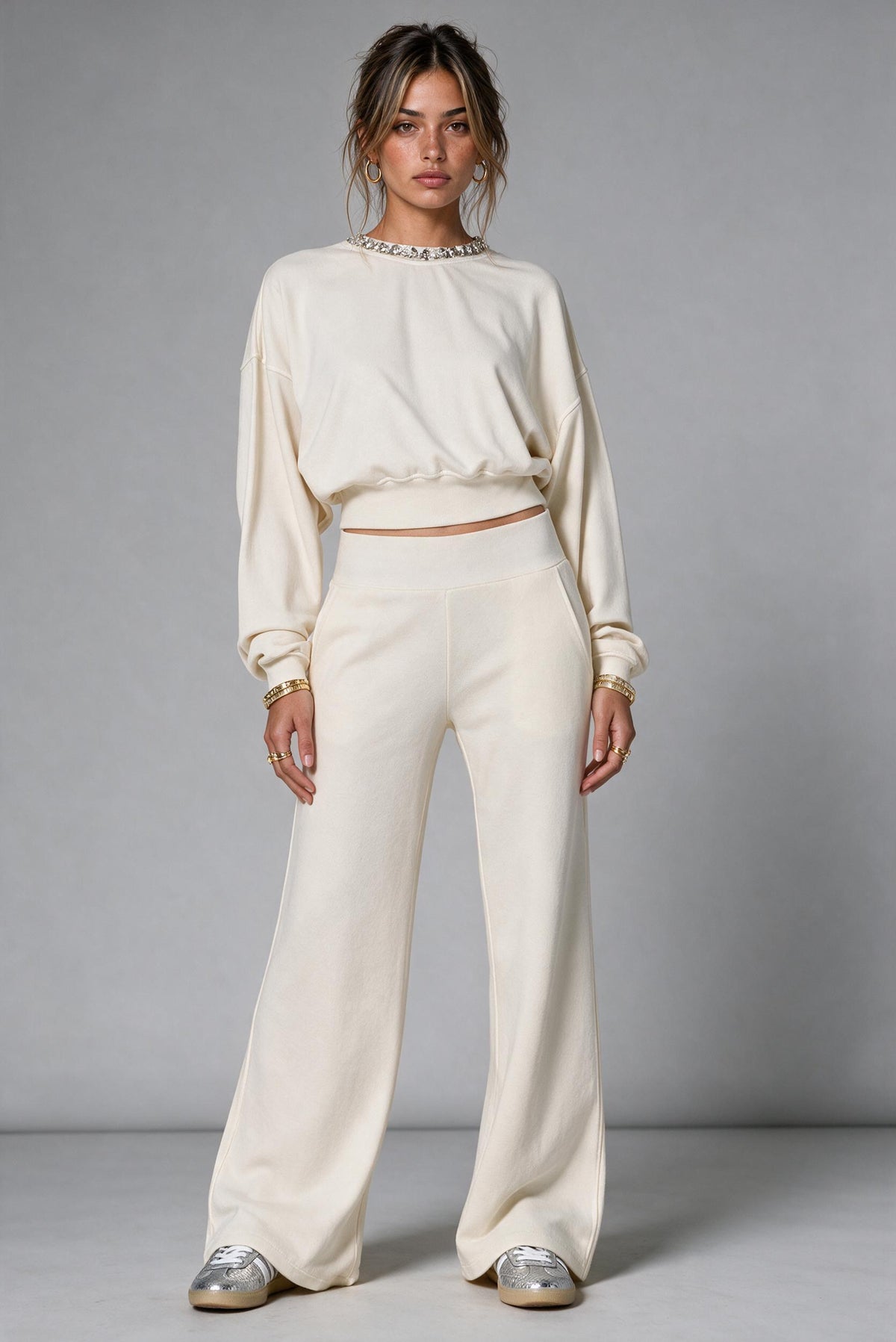A model stands against a neutral gray studio background wearing a matching cream-colored two-piece lounge set consisting of a cropped long-sleeve top with a jeweled neckline and wide-leg trousers, paired with metallic silver shoes.